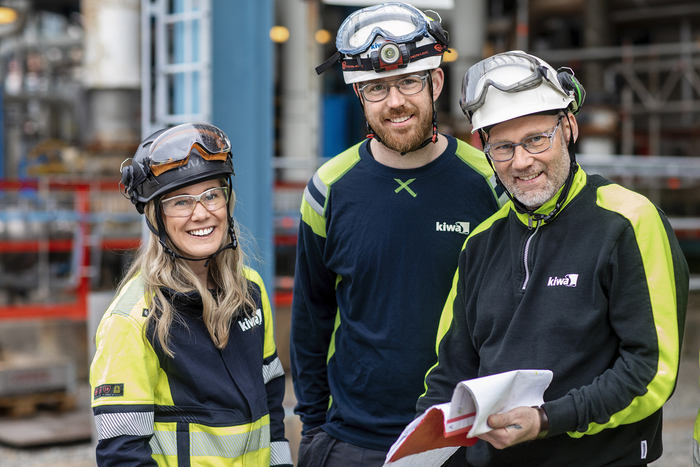 Three Kiwa auditors workers wearing safety gear, including helmets and reflective clothing, standing in an industrial setting with machinery in the background