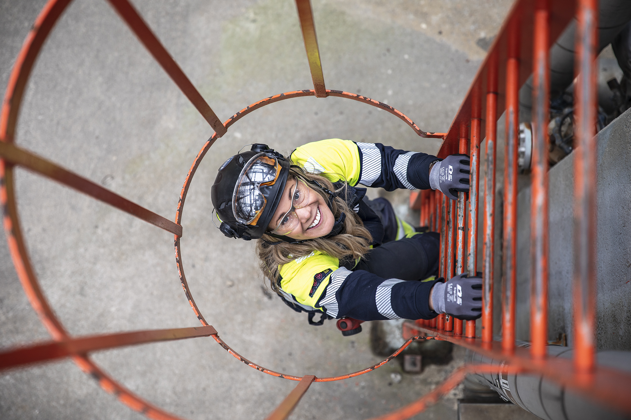 Female Kiwa inspector climbing an industrial ladder during an audit, wearing safety gear and a helmet, smiling up at the camera